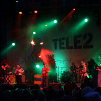 A band performing on a stage under colorful lights, featuring red and green hues, reminiscent of Positivus. Six musicians play various instruments, including guitars, a keyboard, and drums. A large backdrop with the "TELE2" logo and abstract designs is visible behind them. An audience is in the foreground.