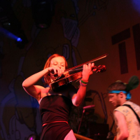 At Positivus, a woman passionately plays the violin on stage, dressed in a black and pink outfit. Behind her, a man with a blue headband plays a guitar. The background, featuring colorful artwork from AB 2008, is lit with vibrant stage lights, creating an energetic atmosphere.