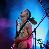 A person stands on stage under vibrant blue and purple lighting at Positivus 2008, playing an electric guitar. They are wearing a sleeveless shirt and a headband with a large feather attached. The person appears enthusiastic, looking upwards with an open mouth, and there are microphones and plants in the background.