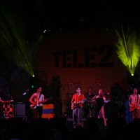 A band is performing on stage under green and yellow spotlights at Positivus 2008. The background features a "TELE2" banner. Musicians are playing keyboards, guitars, and drums with one draped in a colorful flag. The stage, adorned with plants, creates an atmospheric setting for the event.
