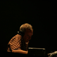 At Positivus 2008, a person with headphones stands at a DJ table in a dark setting. They are wearing a patterned shirt and appear focused on the equipment in front of them, which includes a laptop and other DJ gear. The background is dark, emphasizing the person and the equipment.