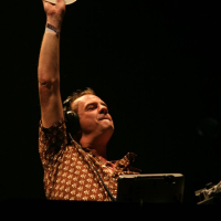 A DJ with headphones holds up a white vinyl record high in the air at Positivus. He wears a patterned shirt and an expression of triumph and concentration. The background is dark, highlighting the DJ and creating a dramatic atmosphere. There is DJ equipment in front of him, reminiscent of AB 2008 energy.