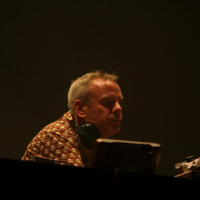 A person with short gray hair and wearing a patterned shirt is sitting in front of electronic equipment, possibly DJing at Positivus. They have headphones around their neck and appear focused on the task. The background is dark, creating a moody atmosphere with minimal lighting on the person's face.