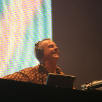 A person smiling broadly while performing as a DJ at Positivus. They are wearing headphones and standing behind a DJ console with a laptop featuring an Apple logo. The background is illuminated with vibrant, colorful lights. The person appears to be enjoying the moment during the performance.