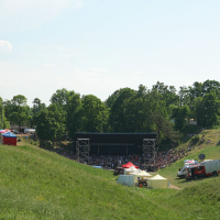 A large outdoor concert stage is set up in a grassy, wooded area. Crowds are gathered in front of the stage, ready for the Punk Laulupidu festival. Several tents and a red truck, possibly emergency services, are positioned to the right of the audience. The sky is clear and sunny, and the surrounding trees are lush and green.