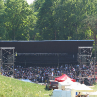 A large crowd gathers in a natural amphitheater with lush green trees in the background for Punk Laulupidu. The audience is seated in front of a stage with a large black backdrop, flanked by metal scaffolding towers. Several tents and vehicles are visible in the foreground, indicating an outdoor punk festival.