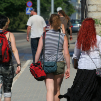 Three people walking down a sunny sidewalk. The person on the left, with short hair and a red backpack, carries a black flag that gives off a punk vibe. The middle person wears a striped tank top and khaki shorts. On the right, someone with long red hair sports a white blouse and black skirt. Trees and a beige building are in the background, reminiscent of Laulupidu gatherings
