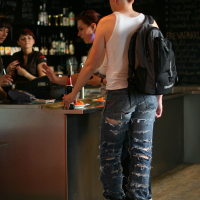 A punk with a mohawk haircut, wearing a white tank top, ripped jeans, black boots, and a black backpack, stands at a bar counter. They are holding a bottle and interacting with one of the three people behind the counter. The bar has shelves with bottles and a chalkboard with handwritten text.
