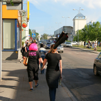 A group of people walk on a sidewalk next to a street, some wearing black punk outfits. One person with a pink wig has text on their back. Another person carries a long, rolled-up item on their shoulder. Cars are parked along the road; buildings and trees line the street under a clear blue sky.