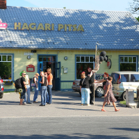 A group of people are gathered in front of a yellow and green building with a sign that reads "Hagari Pitsa." Parked cars are visible nearby. One person, looking punk, is holding black balloons. A person with flowers walks by on the sidewalk. A bright blue sky and greenery form the background.