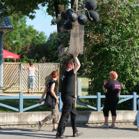 A group of people dressed in black, embodying a punk vibe, stand on a sidewalk near wooden fencing. One person holds balloons and a sign. Another person, with orange hair, takes a photo. Trees and greenery fill the background, and a child sits on the fence. The scene takes place on a sunny Laulupidu day.