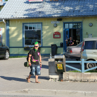A punk sporting a black t-shirt, denim shorts, green mohawk, and neon pink sunglasses strolls down the sidewalk. He's carrying a cylindrical bag and a plastic bottle. Behind him, the two-story yellow building of "Hagari Pub" with its grey tiled roof stands out. A tan car is parked nearby.