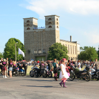 A large gathering of people is taking place outdoors near a historic building with twin towers. Multiple motorcycles are parked, and individuals are socializing. A child in a colorful outfit walks in the foreground, adding to the vibrant ambiance reminiscent of a festive Punk Laulupidu event under clear skies.
