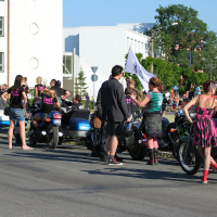 A group of people, mostly wearing black and pink punk-style outfits, gather around motorcycles on a sunny day. Some are sitting on the bikes while others stand nearby. Buildings and trees are in the background, and a few people hold a white flag. The scene has a vibrant, festive Laulupidu atmosphere.