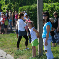 A group of people standing outdoors on a sunny day, gathered along a curb and green hedge, perhaps enjoying the vibrant atmosphere of Laulupidu. In the foreground, a girl in a white top and green skirt stands next to a woman wearing sunglasses and holding a beverage. Other adults and children are spread out along the path.