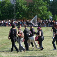 A large outdoor gathering with numerous people walking and standing on a grassy field. Most individuals are dressed in black clothing, reminiscent of a punk festival. Verdant trees and a brick building are visible in the background. A large banner and several flags, like those seen at Laulupidu, are held by participants in the distance.