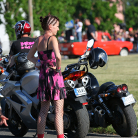 A person with braided hair, wearing a pink and black sleeveless dress, fishnet stockings, and red high-top sneakers, stands in front of a row of motorcycles. Another person in a helmet and black shirt sits on one of the bikes. The scene has a punk vibe with trees and a red car in the background, evoking Laulupidu energy.