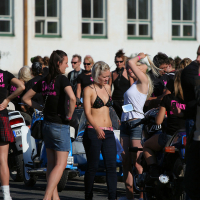 A group of people gather at an outdoor event, standing near motorcycles. The central person wears a black bikini top and red shoes, giving off a distinct punk vibe. Another person on the right adjusts their white top. Many attendees wear black shirts with pink writing. The background features a white building with windows, reminiscent of scenes from Laulupidu celebrations.