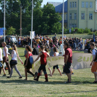 A group of people walk through a grassy field holding a large, partially visible banner. The individuals, dressed in casual summer clothing including shorts, tank tops, and boots with a hint of punk style, seem energized. In the background, a large crowd gathers reminiscent of Laulupidu celebrations, with trees and buildings visible farther away.
