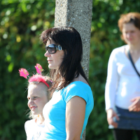 A woman in sunglasses and a blue shirt stands beside a young girl wearing a headband with pink decorations. They are outdoors, leaning against a tree with lush greenery in the background. Other people, possibly gathering for the vibrant Laulupidu festival, appear indistinctly in the background.