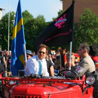 A colorful parade weaves through the streets, with some participants riding a red vehicle. Two prominent figures, one in a white shirt and blue tie, and another in a beige suit, wave flags. Black and Swedish flags flutter alongside an onlooker's small Estonian flag. Trees and buildings frame this festive Laulupidu scene.