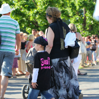 A woman wearing a long, flowing skirt and black top walks hand-in-hand with a child in a black T-shirt and jeans. They’re part of the crowd at Laulupidu, surrounded by greenery and other attendees, some holding bags and pushing strollers. The scene has an eclectic punk vibe woven in with tradition.