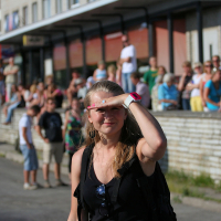 A woman with long hair, wearing a black sleeveless top and sunglasses, is shielding her eyes from the sun with her left hand. She stands on a street in front of a crowd gathered along a brick wall and sidewalk, giving off a punk vibe. The surrounding area includes buildings and parked cars.