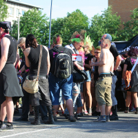 A group of people are gathered outdoors during the daytime at what appears to be a lively Laulupidu. They display various styles, including colorful punk hairstyles, black clothing, and some with bare torsos. The background features trees and a few buildings. A couple of banners are held up among the crowd.