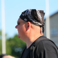 A person wearing sunglasses, a black bandana with a white pattern, and a black shirt stands turned to the side against a backdrop of greenery and a blurred building. With light skin and short hair, they carry a black bag adorned with straps and buttons, exuding an edgy punk vibe reminiscent of Laulupidu.