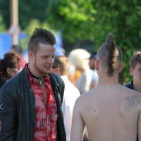 Three men with punk hairstyles and attire are conversing outdoors. The man on the left wears a black jacket with patches and a red shirt, while the man in the middle is shirtless, revealing a back tattoo. A third man stands in the background, wearing a black shirt. The setting resembles Laulupidu festivities in a green park area.