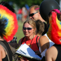 A group of people gather outdoors, many sporting vibrant, punk-inspired rainbow-colored mohawk wigs. A woman at the center, wearing sunglasses and a red shirt, holds papers and a clipboard. The background has trees and a sunny atmosphere, reminiscent of Laulupidu's bright and energetic setting.
