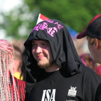 A man with a beard wearing a black shirt with text and logos, and a black shirt draped over his head, stands outside on a sunny Laulupidu day. Other people are visible in the background, with one person having brightly colored punk braids. Trees and a blurred crowd can be seen in the background.