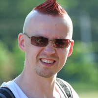 A person with a red mohawk hairstyle, wearing sunglasses, a white tank top, and a black backpack, is seen outdoors on a sunny day. They are smiling broadly, revealing their teeth, with a green, blurred natural background—a true punk spirit enjoying the sunshine.