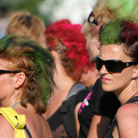 A group of people with colorful, punk-style mohawk hairstyles are standing together outdoors at Laulupidu. Two prominent individuals wear black sunglasses; one has green-tipped hair and the other has green and red hair. The background is filled with more people sporting vibrantly colored, creative hairdos.