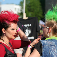 Two women with brightly colored mohawks, one red and one green, are engaged in conversation outdoors. The woman with the red mohawk wears a punk-inspired red top and black vest, while the woman with the green mohawk sports a denim vest and lime green scarf. Both are wearing sunglasses.