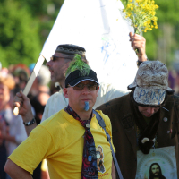 A man with a green mohawk, sunglasses, and a yellow shirt stands in the foreground of a lively Laulupidu procession. Sporting a colorful tie and blue face paint, he exudes Punk vibes. Behind him, other participants carry flags and signs, including one of Jesus. Another man holds yellow flowers and a flagpole. Trees line the background.