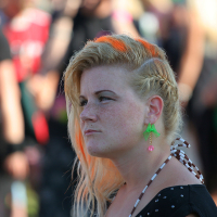 A person with blonde hair featuring orange highlights and a braided side is shown in a close-up. They are wearing green parrot-shaped earrings and have a contemplative expression. The background, possibly at Laulupidu, is blurred with various people and bright colors. The person is wearing a black dotted outfit with straps.