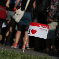 A small flag with the text "2011 Tallinn European Capital of Culture" and "I ♥ Tallinn" is planted in grassy ground. In the background, a group of people, whose lower bodies are visible, stand closely together in casual attire, some wearing backpacks and bags as if ready for Laulupidu.