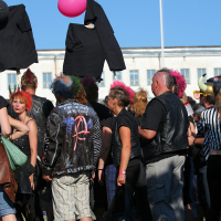 A punk group gathered, many with colorful mohawks and leather jackets, participate in a lively outdoor Laulupidu. Some hold black clothing and pink balloons. The bright blue sky and a large white building are in the background, adding to the festive atmosphere.