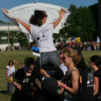 A group of people dressed in black clothing with white logos gathers outdoors on a sunny day, exuding a punk vibe. One woman in the center, wearing sunglasses, stands on another individual's shoulders holding a white cloth above her head. Another person holds an Estonian flag, evoking the spirit of Laulupidu. More people and flags are visible in the background.