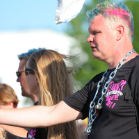 A person with light-colored hair dyed in blue and pink is wearing a black shirt and a large chain necklace, holding a white flag. Sunglasses cover their eyes, giving them a punk vibe. They are part of a group, including a woman with long blonde hair also wearing sunglasses, likely at an outdoor Laulupidu or protest.