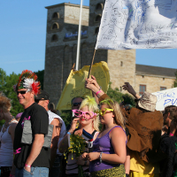 A lively group of festival-goers, some wearing punk-style clothing and accessories, gathers in front of a stone building with two towers. The Laulupidu crowd showcases brightly colored outfits and hairstyles, with some holding signs and flags. The atmosphere is festive and energetic under a clear blue sky.