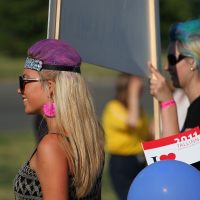 A woman with purple hair and a headband stands outdoors, holding a sign. She wears large pink earrings and sunglasses. Nearby, another punk with colorful hair holds a "Tallinn 2011" sign and a blue balloon. Blurred individuals in the background are in a park-like setting, celebrating Laulupidu.