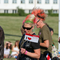 A group of people with colorful punk hair, including neon green and bright orange, stand outdoors holding small flags that read "I ❤️ TALLINN 2011." They're dressed in casual clothing. The background includes a grassy area and a white building. Some seated people are visible in the distance, suggesting a Laulupidu celebration.