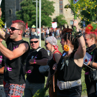 A vibrant group of people, many with dyed hair and colorful clothing, engage in an outdoor celebration or protest. They hold flowers, flags, and signs while smiling and interacting like a punk-themed Laulupidu. The background reveals a crowd and part of a building, indicating a lively public event.