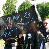 A group of people in punk attire holding a large black banner that reads "PUNK REIR ANKERE 2011 SURE" with various symbols, including an anarchy sign. The individuals, flaunting leather jackets and sunglasses, evoke the spirit of Laulupidu. Trees and a building are visible in the background on a sunny day.