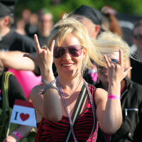 A blonde woman wearing sunglasses gives rock-and-roll hand signs with both hands, capturing the punk spirit. She is smiling and standing in a crowd of people, some of whom are also dressed in black and pink. The crowd appears to be outdoors at Laulupidu, with the woman rocking a striped black and pink top.