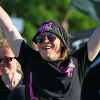 A person with long hair, wearing sunglasses and a bandana, smiles broadly with arms raised in celebration at Laulupidu. They sport a punk-inspired black T-shirt with pink and white graphics. Another smiling individual in sunglasses is partially visible behind them. The background is blurred green foliage and people.