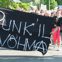 A group of people march outdoors on a sunny day holding a large black banner with white, spray-painted text reading "PUNK IL VÕHMA." The participants, dressed in colorful punk attire with vibrant hair and accessories, embody the spirit of Punk Laulupidu. Trees and other marchers are visible in the background.