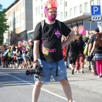 A person stands in the middle of the street holding a camera. They sport a colorful mohawk, face paint, and wear a black t-shirt with pink designs, denim shorts, and sandals. A crowd of people and buildings line the background on this sunny day at Laulupidu, capturing the punk spirit perfectly.